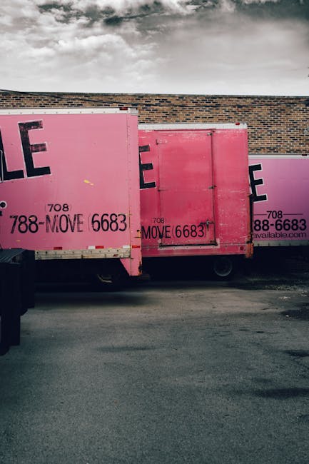 Two pink moving containers with large black and purple lettering are positioned side by side against a brick wall, on a paved outdoor area under a cloudy sky. The containers are used for home relocation purposes, and the visible text includes phone numbers and the word 'MOVE.' One container appears slightly larger, and both are made of sturdy material with metal edges and wheels for mobility. The setting indicates a loading area where items such as cardboard boxes, furniture, and packing materials are transported for moving services. The containers are part of the logistics process managed by Removal Van Finsbury Park, supporting furniture transport and packing during house removals, with some presence of protective coverings or plastic wrap on stored items inside or around the containers. The overall scene reflects a typical moving or packing operation in progress, with no additional personnel or equipment visible.