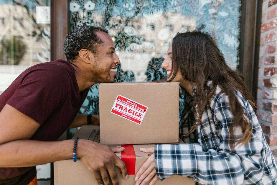 Two individuals, a man and a woman, are engaged in a home relocation process outside a building with a brick wall and a large window in the background. The man, with short dark hair and wearing a maroon t-shirt, is smiling and facing the woman as they share a close moment. They are holding several cardboard boxes together; one box is marked with a red 'Fragile' sticker indicating careful handling, and the boxes are sealed with red tape. The woman, with long brown hair and wearing a blue and white checkered shirt, is also smiling. They appear to be in the midst of packing or unpacking, with the boxes clearly visible in the frame, suggesting a packing and moving activity. The scene is well-lit and captures the collaborative effort involved in a furniture transport or moving process, typical of services provided by Removal Van Finsbury Park for house removals and relocation logistics.