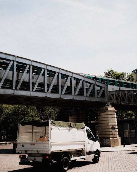 A white delivery van operated by Removal Van Finsbury Park is parked on a paved street beneath a large railway bridge. The van's rear cargo area is partially covered with a green tarp secured with straps, and there are cardboard boxes and packing materials inside, suggesting ongoing packing or loading activities related to home relocation. The van is positioned near the sidewalk, with the loading area accessible for furniture transport or moving boxes. The railway bridge above is constructed of metal trusses supported by stone pillars, with a train visible on the tracks. Surrounding trees and a building are visible in the background, with natural daylight illuminating the scene, emphasizing the logistics involved in a same-day house removal service. Removal Van Finsbury Park’s vehicle is ready for further loading or transport, reflecting professional furniture transport and packing procedures typical of removals services.