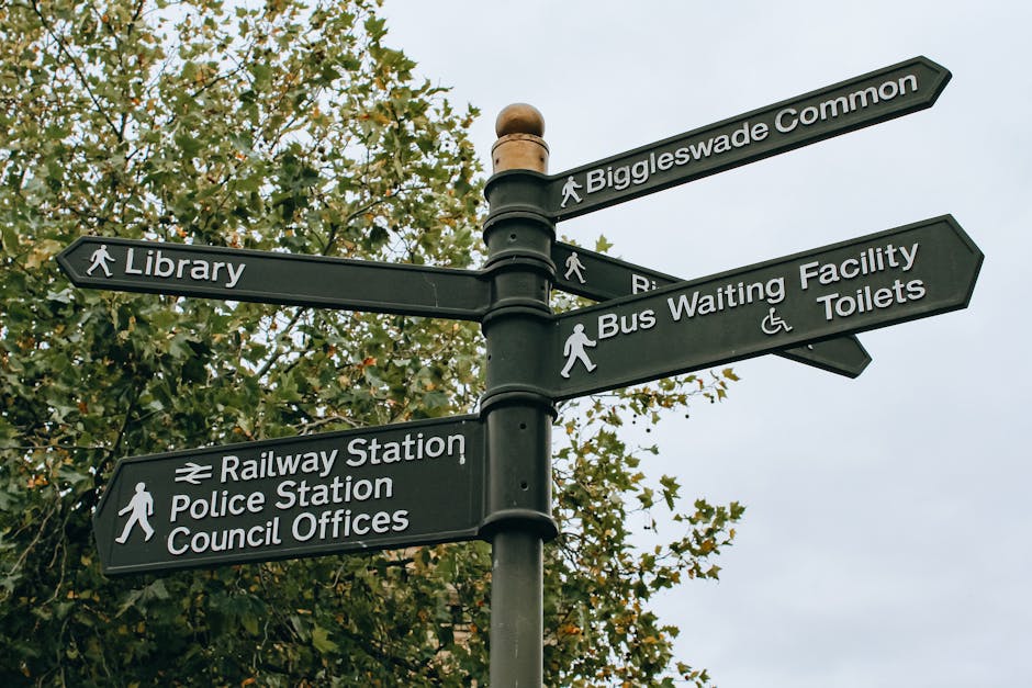 A black multi-directional street signpost located outdoors against a background of green leafy trees and an overcast sky, with six signs pointing in different directions indicating nearby locations such as Biggleswade Common, Library, Railway Station, Police Station, Council Offices, Bus Waiting Facility, and Toilets; the signs feature white text and symbols, including walking figures, a train icon, and accessibility icons. This signage is situated near a property where a home relocation or furniture transport process by Removal Van Finsbury Park could involve navigating close to local amenities and transportation hubs, with the scene capturing elements relevant to urban moving logistics and the organization of relocation routes.