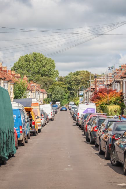 A residential street scene during daytime shows a row of parked cars on both sides of the asphalt road, with various makes and models, including small hatchbacks and larger vans. Toward the far end of the street, a blue and white Removal Van Finsbury Park is visible, positioned for loading or unloading. The van's rear doors are open, revealing the interior with packed cardboard boxes and bubble wrap, indicating a home relocation process. In front of the van, a large wooden furniture dolly is being used to transport a partially visible piece of furniture wrapped in protective fabric and secured with plastic straps. The street is lined with terraced houses featuring brick facades, some with small garden areas or hedges, and there are overhead electrical wires stretching across the scene. The background includes lush green trees and a partly cloudy sky. This image captures the logistics of furniture transport and packing in a typical residential moving scenario, illustrating the work involved in home removals and the use of professional services like [COMPANY_NAME].