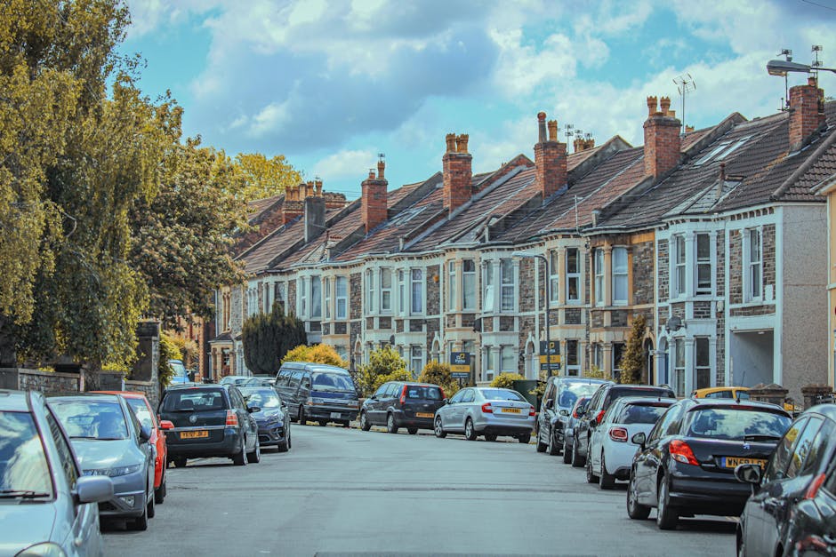 A residential street lined with terraced houses featuring brick and white painted facades, multiple chimneys, and tiled roofs under a partly cloudy sky during daytime. Parked cars, including sedans and hatchbacks, occupy both sides of the street, with some close to the pavement and others slightly set back. Leafy trees provide some greenery along the sidewalk. Inside one of the houses, visible through a doorway or window, there are cardboard boxes, wrapped furniture, and packing materials indicating a home relocation process. The scene suggests an active moving day with furniture and boxes prepared for loading onto a van, supported by the presence of a removal van from Removal Van Finsbury Park nearby, possibly parked on or adjacent to the street. This setting exemplifies typical house moving logistics, with the focus on packing, furniture transport, and loading procedures.